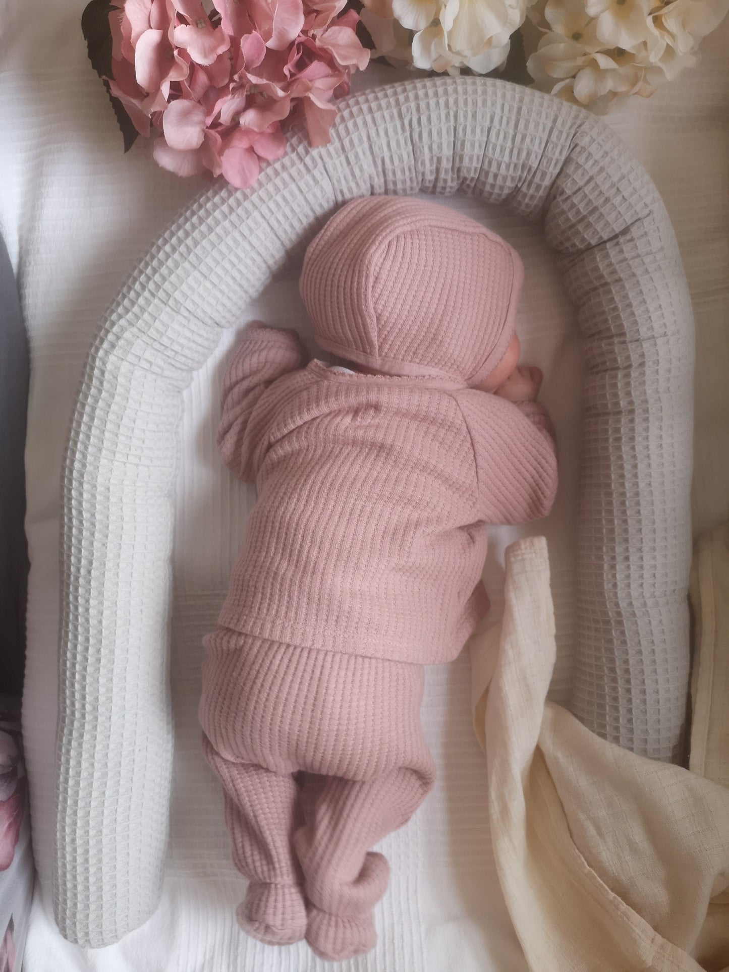 Baby in rosa Outfit liegt geborgen im grauen Waffelpiqué-Nestchen mit Blumen – perfekte Geschenkidee