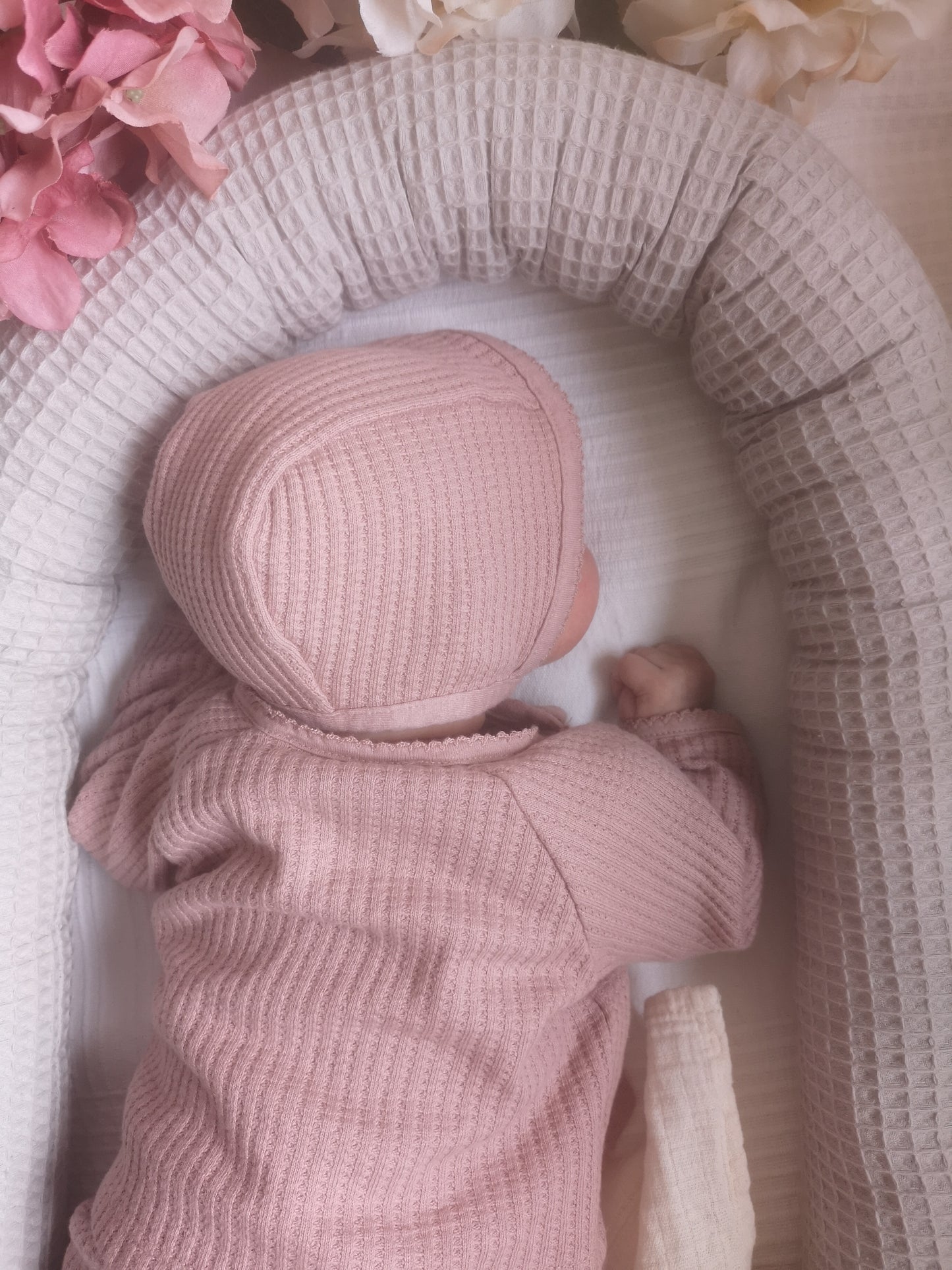 Neugeborenes in rosa Kleidung schläft sicher im grauen Waffelpiqué-Babynestchen mit Blumen-Deko