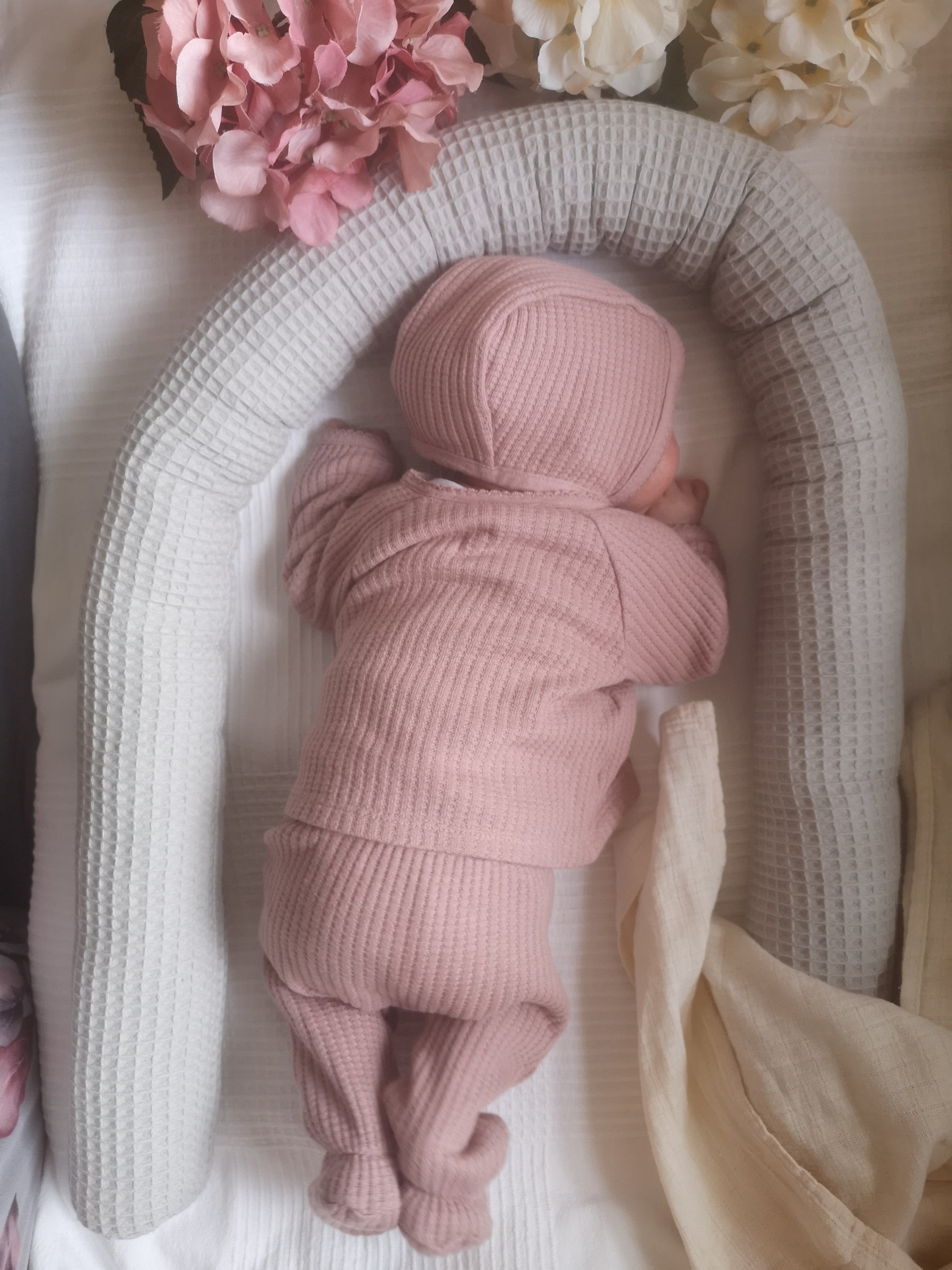 Baby in rosa Outfit liegt geborgen im grauen Waffelpiqué-Nestchen mit Blumen – perfekte Geschenkidee