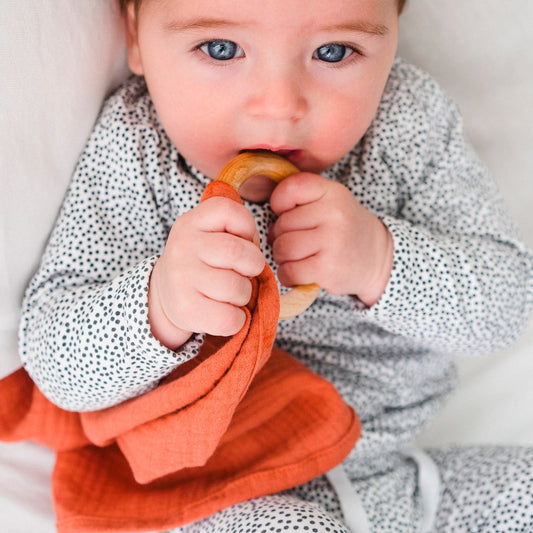 Baby beißt auf ein orangefarbenes Schmusetuch mit Holzring – ideal ab 3 Monaten zum Greifen und Zahnen